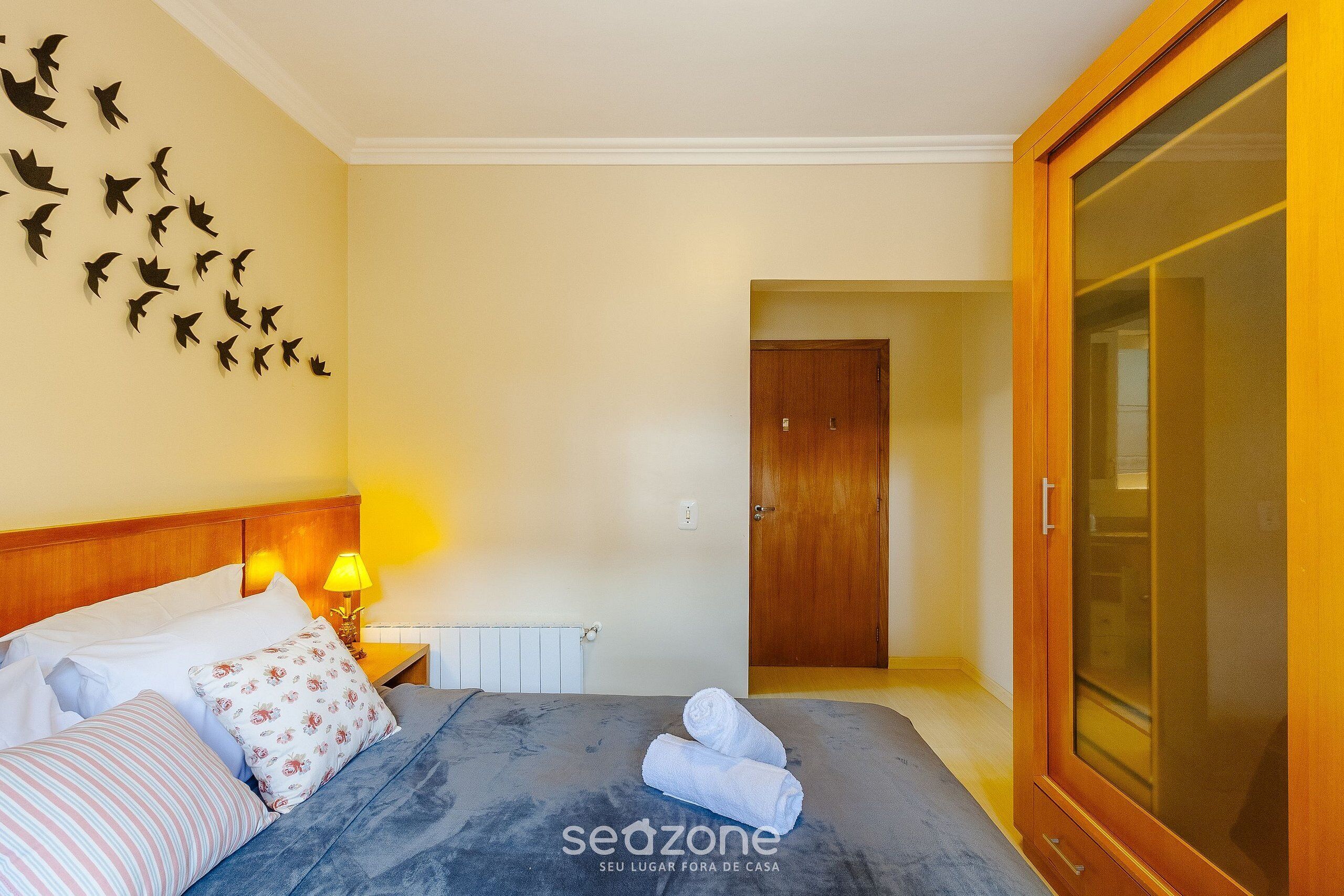 A hotel room with a bed, pillows, a nightstand with a lamp, a wardrobe, and a wall with bird cutouts.