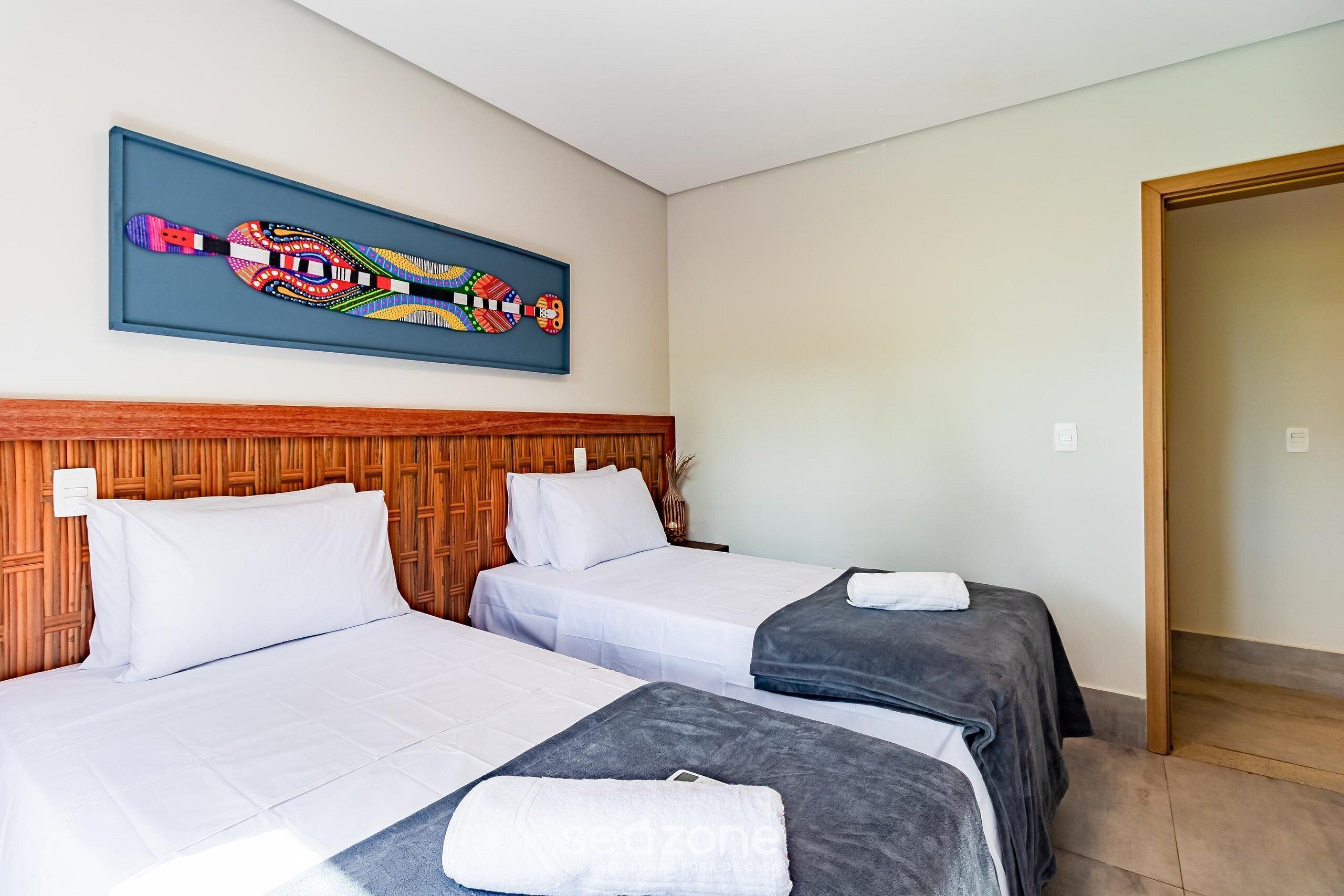 A hotel room with two beds, a wooden headboard, a framed artwork above the beds, and a door to the right.