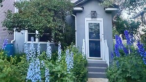 Una casa de dos pisos con un porche delantero, un pequeño jardín con flores azules y un buzón.