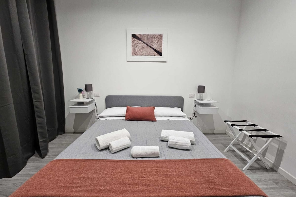 Una habitación de hotel moderna con una cama, dos mesitas de noche, un velador, una obra de arte grande en la pared y una ventana con cortinas.
