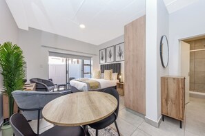 A modern hotel room with a bed, a dining area with a round table and chairs, a large potted plant, and a wardrobe.