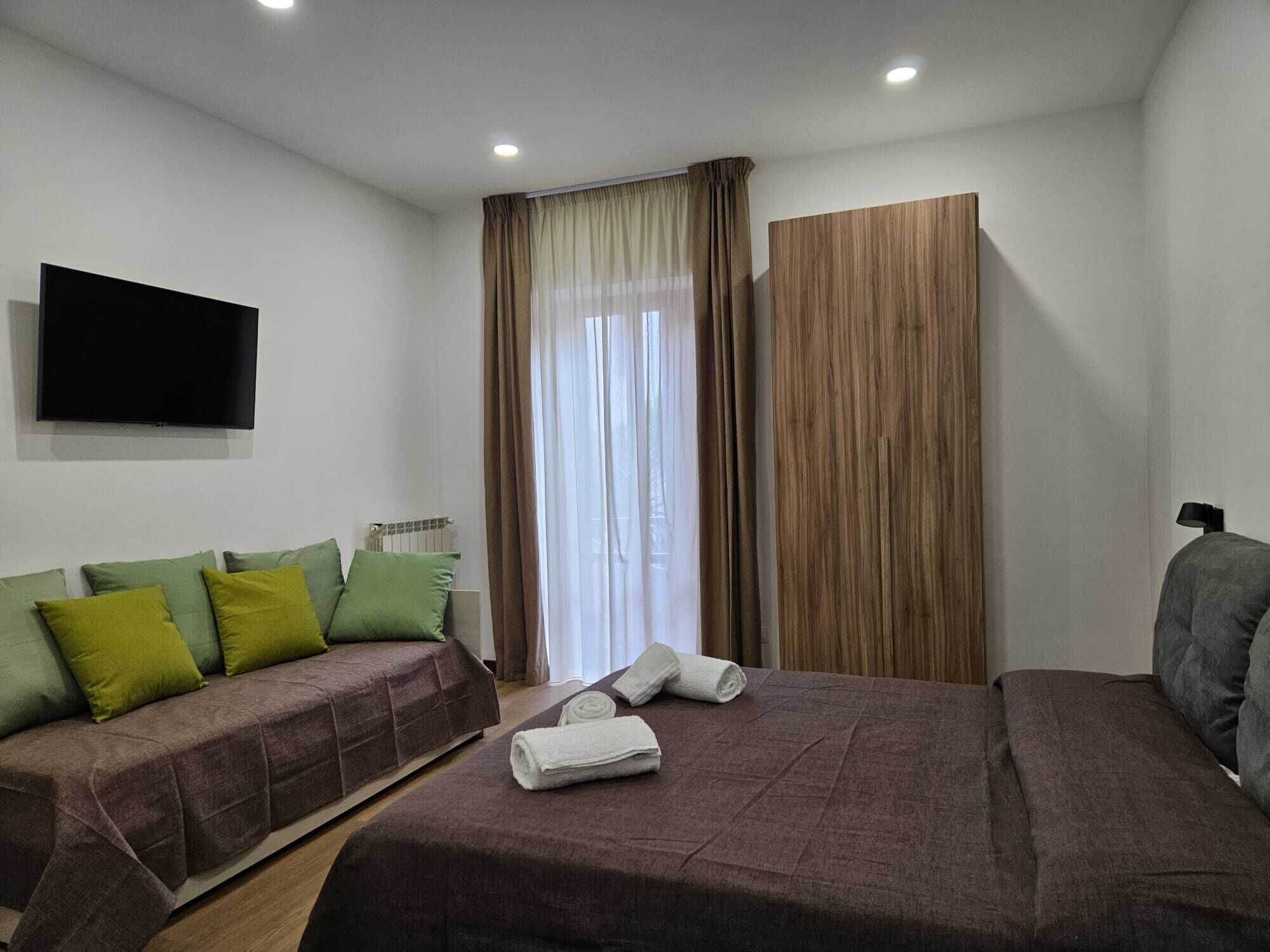 Una habitación de hotel con cama, sofá, televisión y ventana con cortinas.