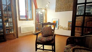 A cozy living room with a wooden chair, a cushioned stool, and a wicker chair. A large window with curtains is visible, and a mirror hangs on the wall. - Loft in the heart of Nice (Nice)