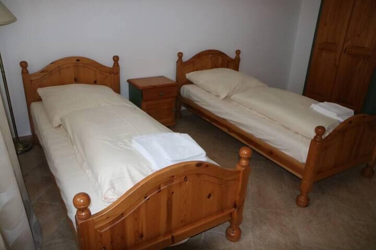 Two wooden single beds with nightstands, a wooden wardrobe, and a lamp in a room with plain walls.