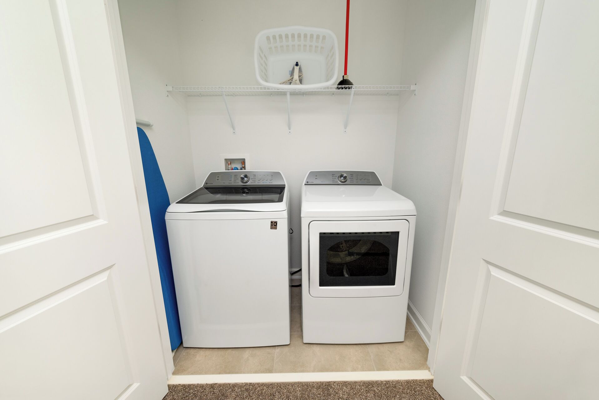 A laundry room with two white washing machines and a dryer, a white laundry basket on a shelf, and a blue towel on the floor.