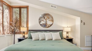 A bedroom with a large bed, two bedside tables with lamps, a decorative tree wall art, and a window with sheer curtains.