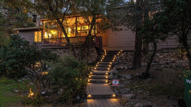 A wooden staircase leading to a house with a porch, surrounded by trees and a stone wall.