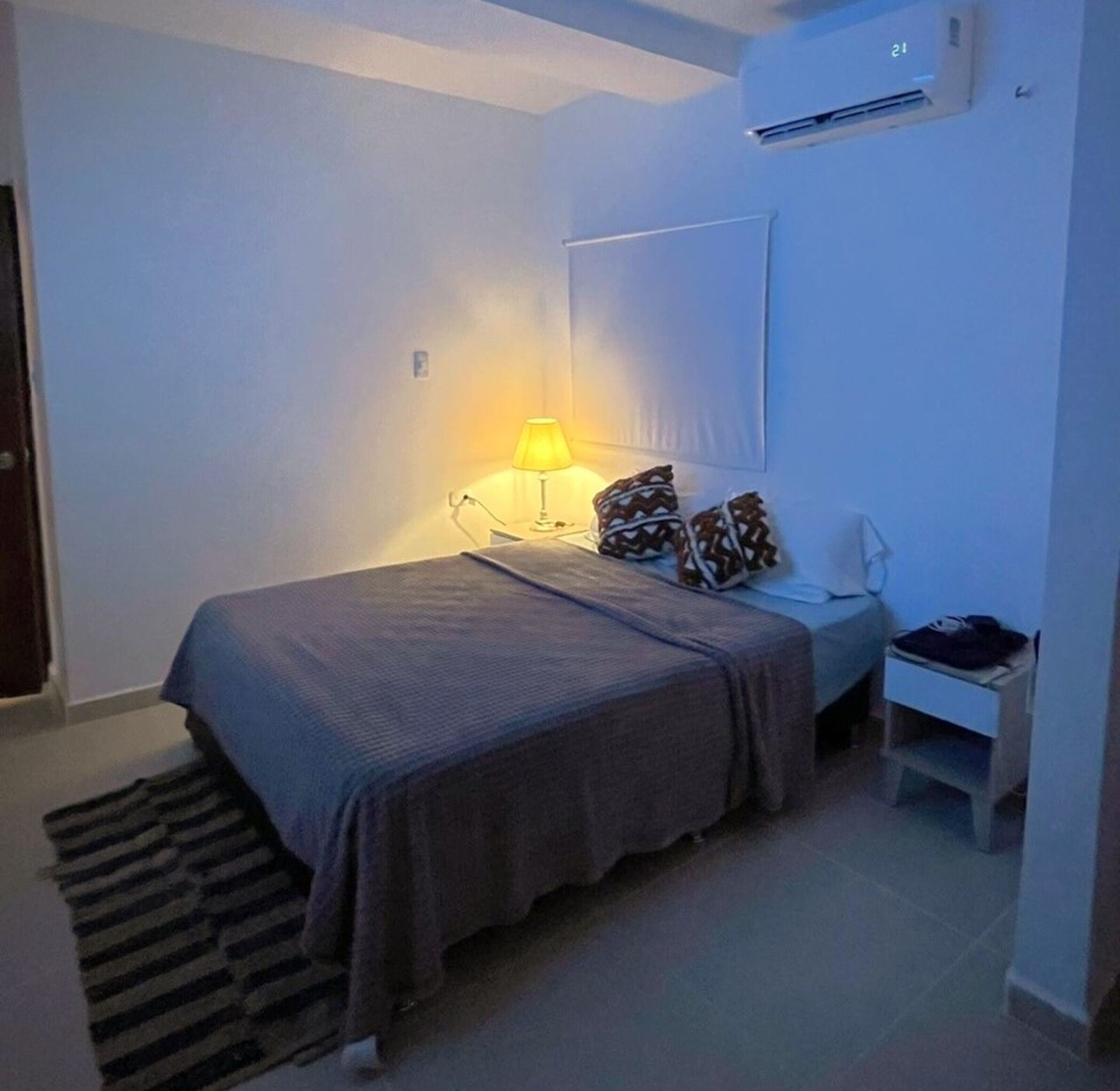 Un dormitorio pequeño y ordenado con cama, mesita de noche, lámpara y unidad de aire acondicionado.