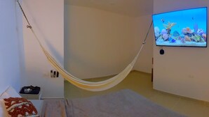 A room with a hammock, a bed, a television displaying an underwater scene, and a small table with a bag and a red item.