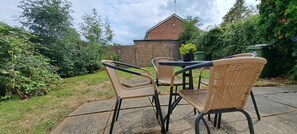 Een terras buiten met rieten stoelen en een tafel, omgeven door een bakstenen muur en groen.