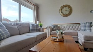 A living room with two sofas, a wooden coffee table, a wall clock, and a window with curtains. - Generous 3 bed in the heart of the village (Sawtry)
