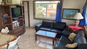 Una sala de estar con un sofá de cuero negro, una mesa de centro, un televisor sobre un mueble de madera, una mesa de comedor con sillas y una ventana con cortinas.