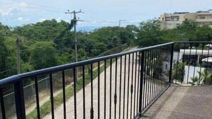 Un balcon doté d’une rambarde en métal, donnant sur une route bordée de verdure et avec des bâtiments en arrière-plan.