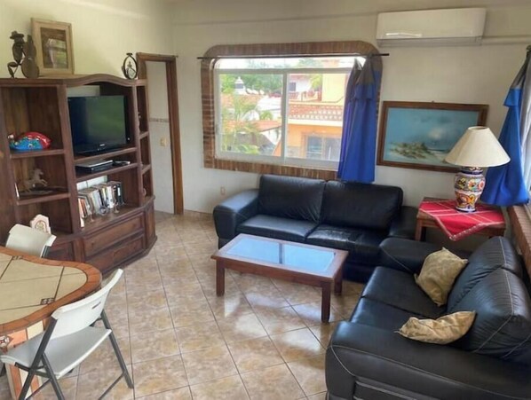 Una sala de estar con un sofá de cuero negro, una mesa de centro, un televisor sobre un mueble de madera, una mesa de comedor con sillas y una ventana con cortinas.