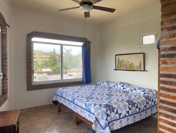 Un dormitorio con cama, ventilador de techo, ventana con cortinas e imagen enmarcada en la pared.