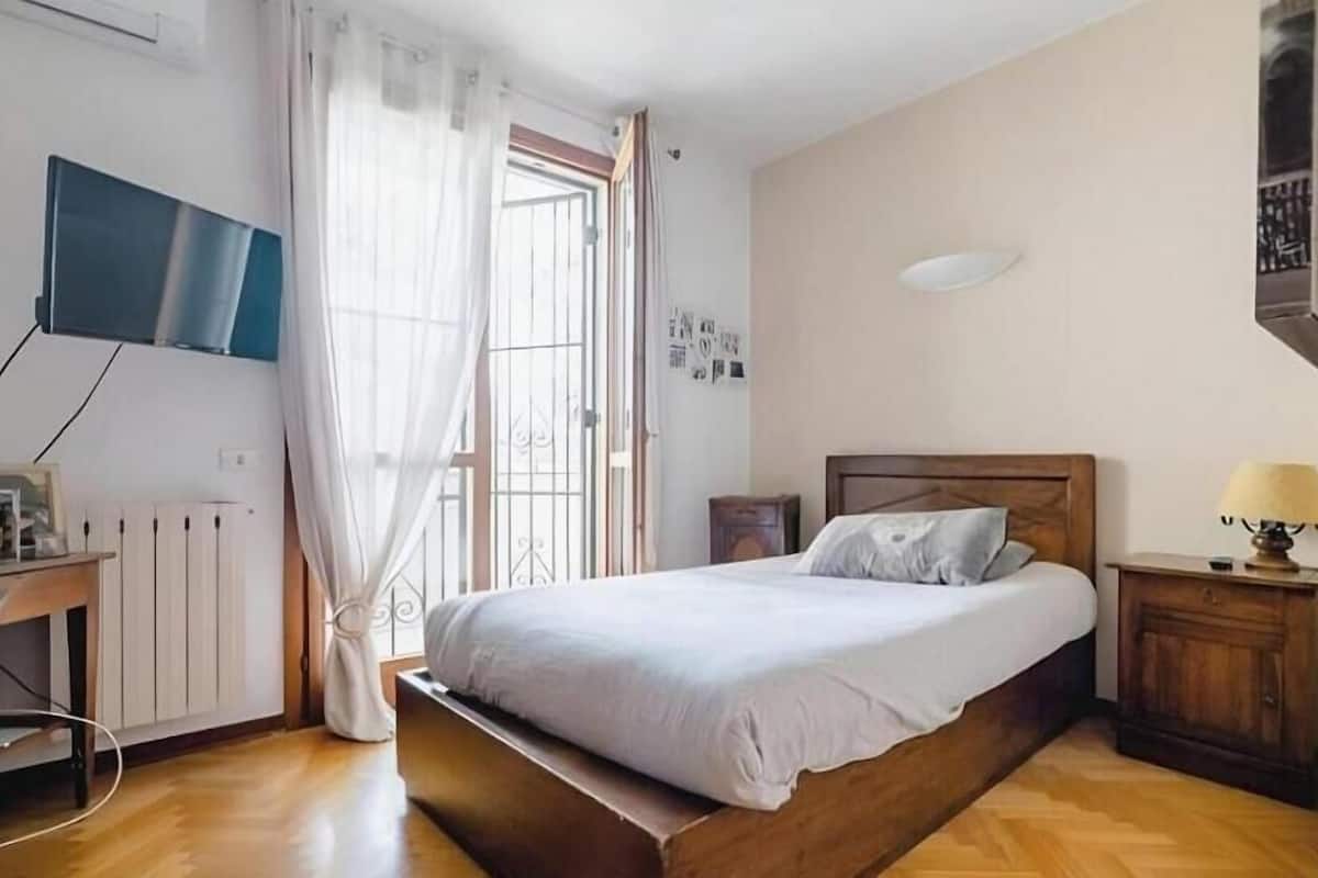A bedroom with a wooden bed, a desk with a computer, a TV mounted on the wall, and a window with curtains.