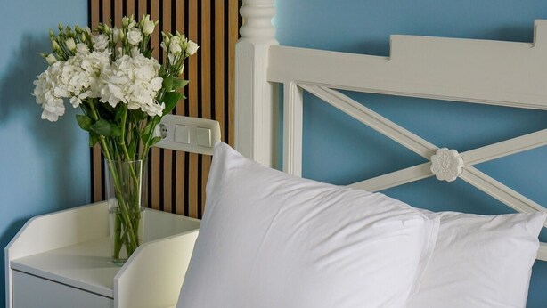 A bed with white bedding and pillows, a bedside table with a vase of white flowers, and a white headboard with a decorative element. - NUEVO HOTEL PARQUE CABAÑEROS (Horcajo de los Montes)