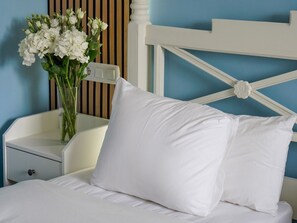 Una cama con ropa de cama y almohadas blancas, una mesita de noche con un florero de flores blancas y un cabecero blanco con un detalle decorativo.
