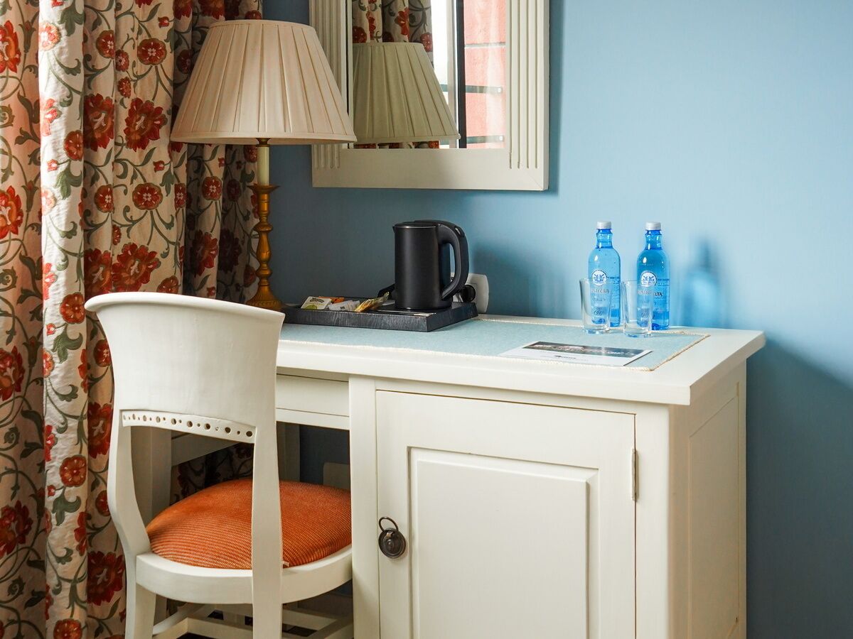 Ein kleines Zimmer mit einem weißen Schreibtisch, einem Sessel mit orangefarbenem Kissen, einer Lampe, einem Wasserkocher und zwei Wasserflaschen.