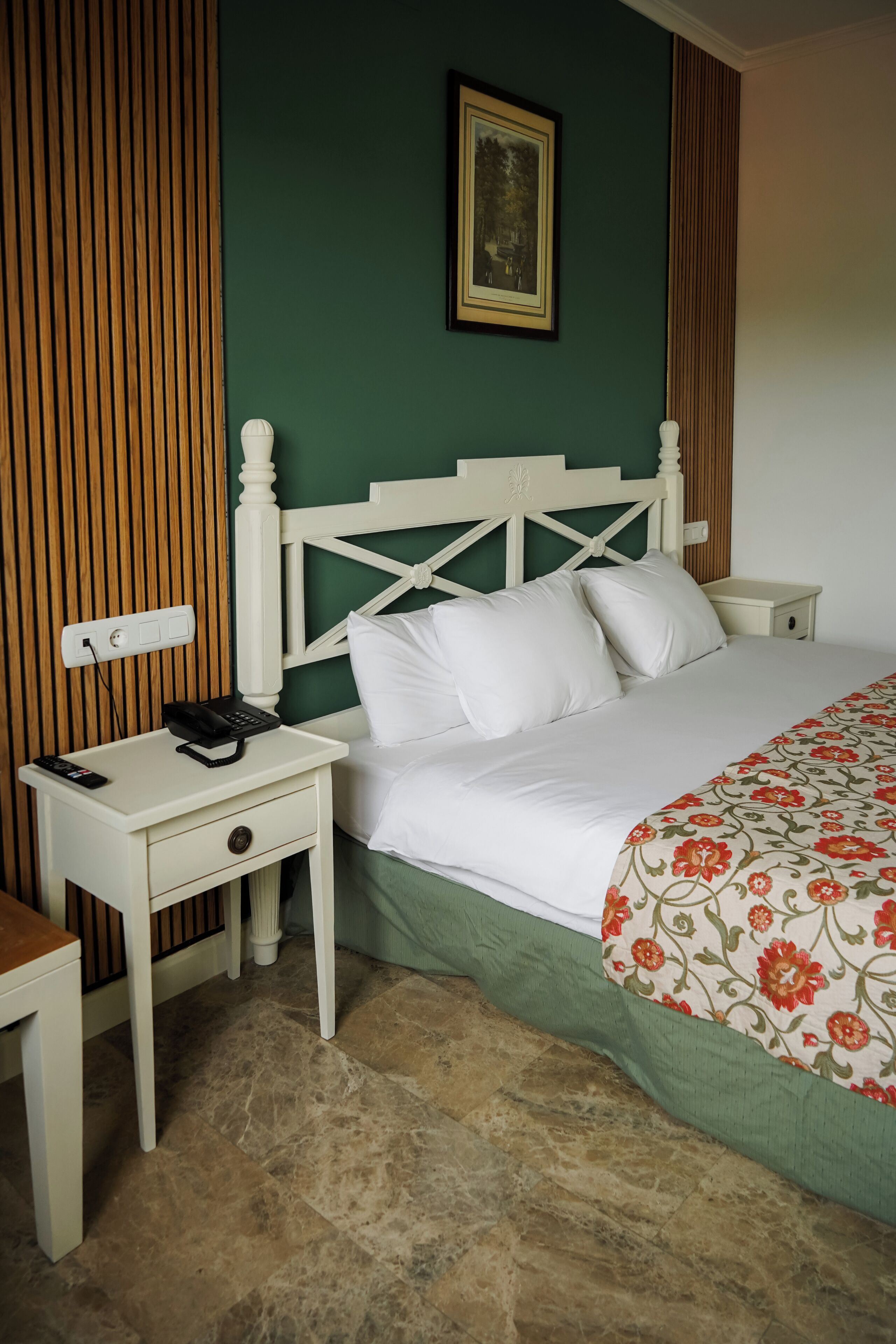 Una cama bien hecha con ropa blanca, un cabecero de madera, una mesita de noche con un teléfono y una pared verde con un cuadro enmarcado.