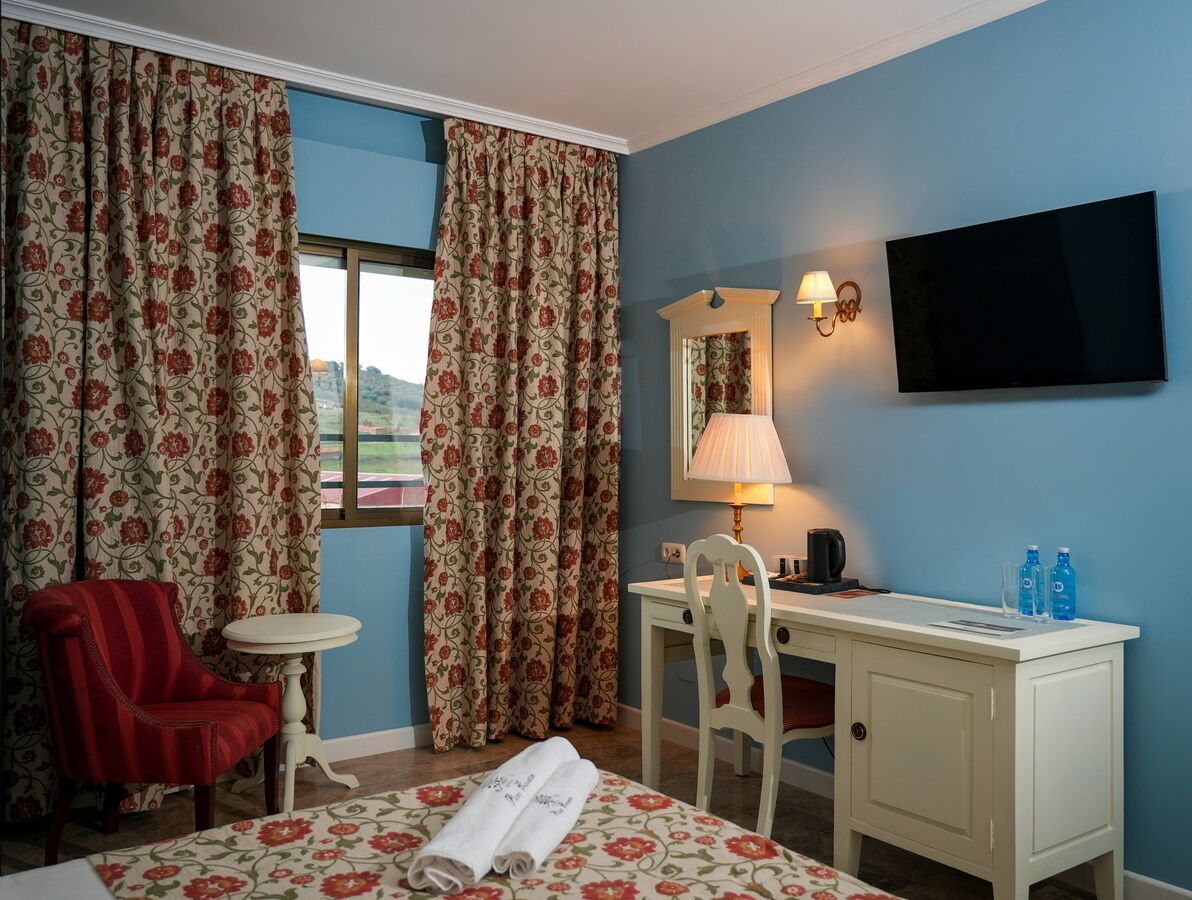 A hotel room with a bed, two chairs, a desk, a television, a lamp, and a window with curtains.