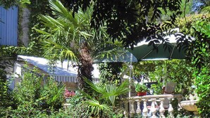 A lush garden with a palm tree, a stone fountain, and a covered seating area with a chair and umbrella. - Spacious 8-bedroom country house in Villepinte with private pool and parking (Villepinte)
