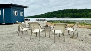 A lakeside seating area with a blue building, outdoor chairs, and a view of a forested hill.