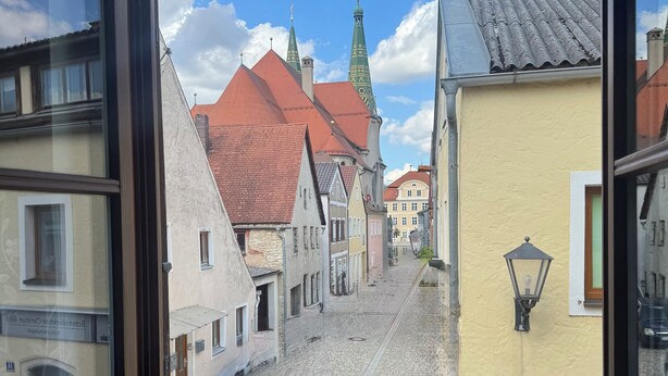 Udsigt gennem et vindue af en brostensbelagt gade med historiske bygninger, heriblandt en kirke med et grønt spir.
