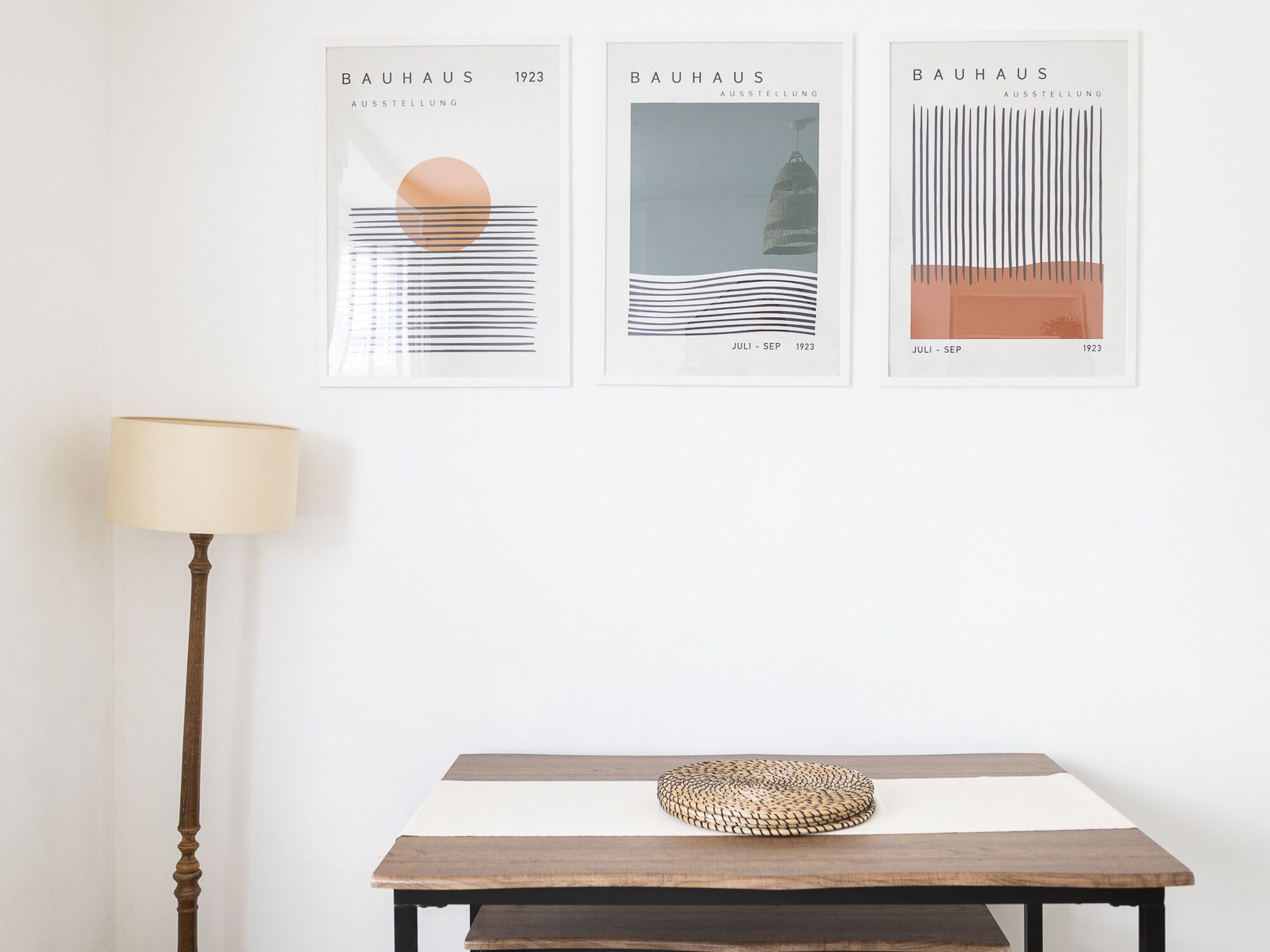 Trois œuvres d’art encadrées, intitulées « BAUHAUS AUSSTELLUNG », datant de 1923, sont exposées sur un mur blanc, au-dessus d’une table en bois sur laquelle se trouve un panier tressé.