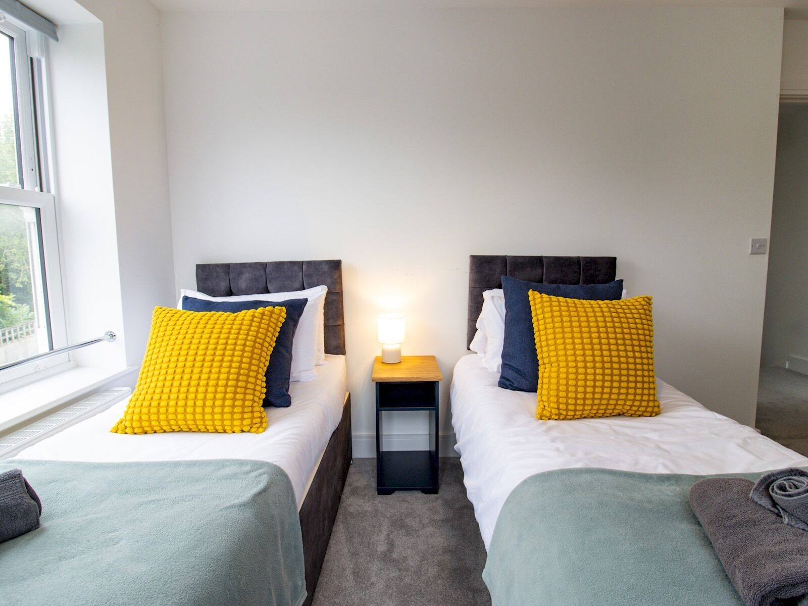 Ein Doppelzimmer mit zwei Betten, jeweils mit Kopfstütze, einem Nachttisch mit Lampe und einem Fenster.