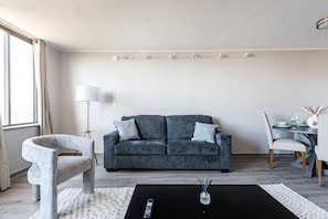 Un salon moderne avec un canapé gris, un fauteuil original, une table à manger avec des chaises, un lampadaire et une table basse noire.