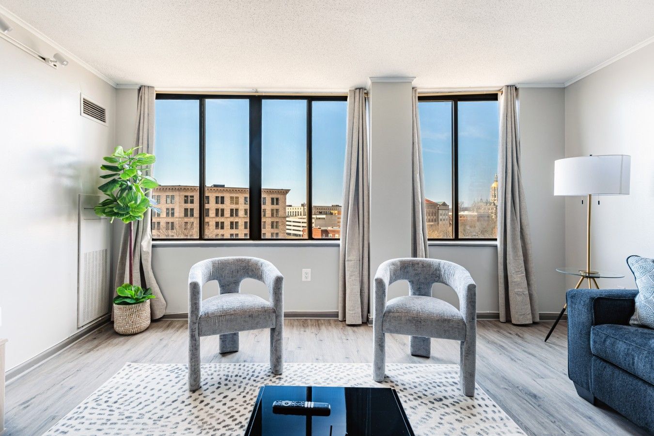 A modern living room with two grey armchairs, a blue sofa, a floor lamp, and a large window with a city view.