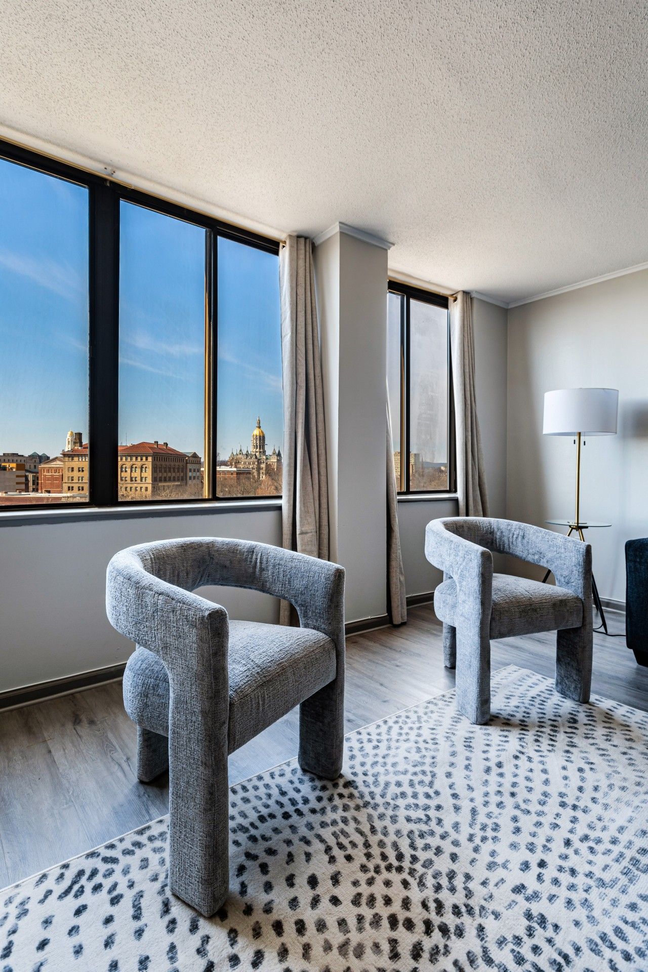Habitación de hotel con dos sillones grises tapizados, una alfombra estampada, una lámpara de pie y vistas de un paisaje urbano a través de amplios ventanales.