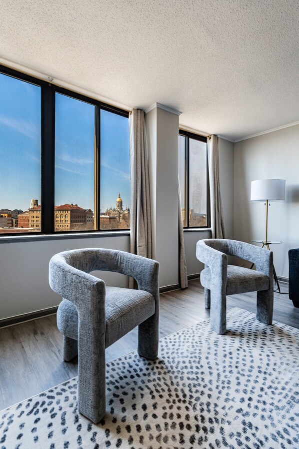 A hotel room with two grey upholstered chairs, a patterned rug, a floor lamp, and a view of a cityscape through large windows.