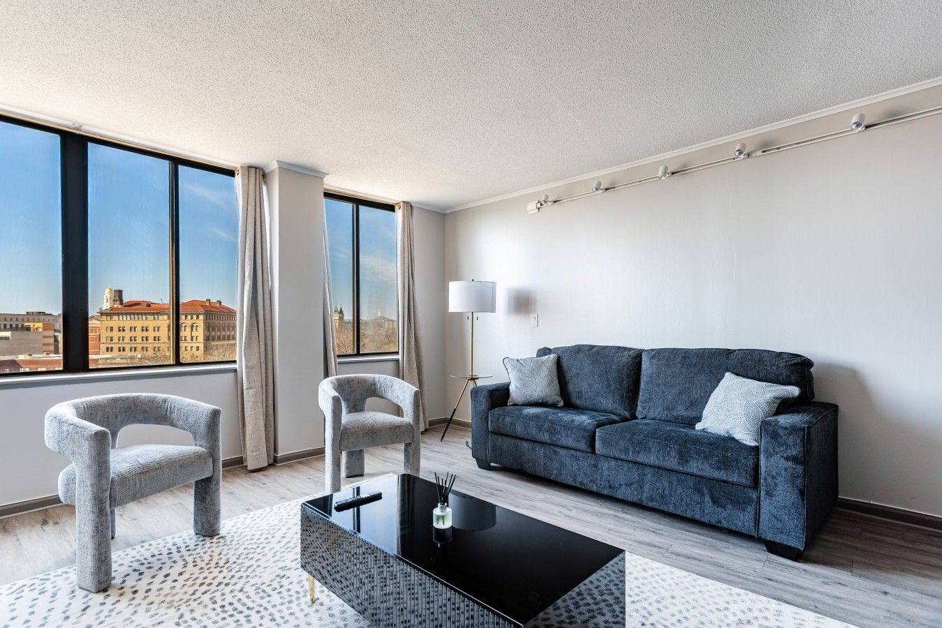 A modern living room with a sofa, two armchairs, a coffee table, and a floor lamp. Large windows offer a city view.