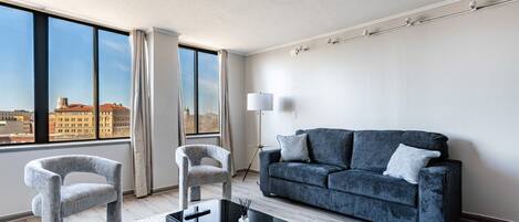 A modern living room with a sofa, two armchairs, a coffee table, and a floor lamp. Large windows offer a city view.
