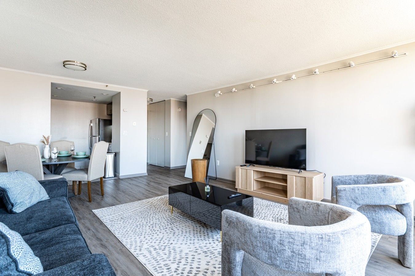 A modern living room with a flat-screen TV, grey upholstered chairs, a coffee table, and a dining area with a table and chairs.