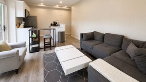 Un salon moderne avec un canapé gris, une table basse blanche et un fauteuil gris. On y trouve également un coin cuisine avec des meubles blancs et une table à manger.