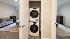 Une buanderie avec des lave-linge et des sèche-linge superposés, un canapé gris et une commode en bois foncé dans un appartement moderne.