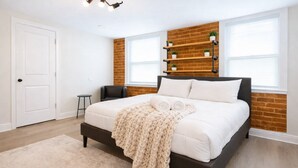 A bedroom with a bed, a chair, a door, a window, and a brick wall.