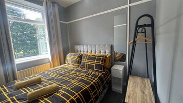 Un dormitorio con cama, radiador, espejo, mesa de madera y ventana con cortinas.