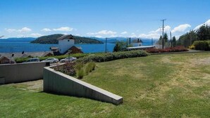 Un espacio verde con un banco de hormigón, un pequeño edificio blanco y vistas a un lago y montañas lejanas.