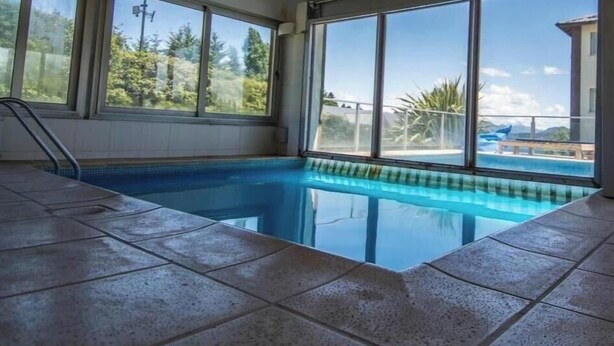 Una piscina interior con amplios ventanales, suelo de baldosas y techo de madera.