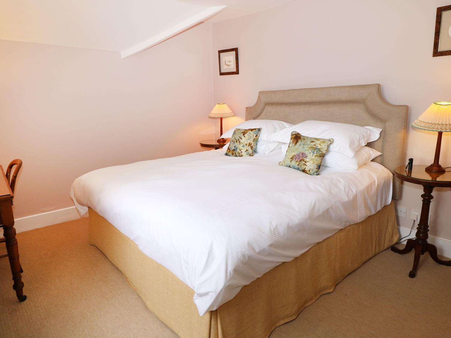 A neatly made bed with white linens and floral pillows, a wooden nightstand with a lamp, and a small table with a lamp.