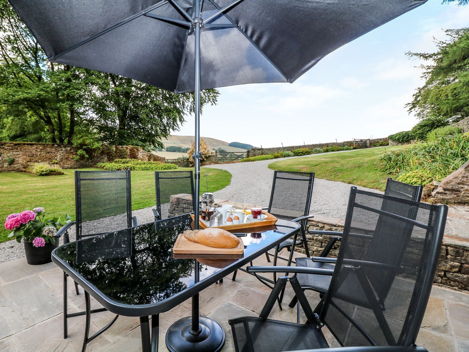 Ein Essbereich im Freien mit Tisch und Stühlen, einem großen Sonnenschirm und einer schönen Aussicht auf einen Weg und Grünflächen.