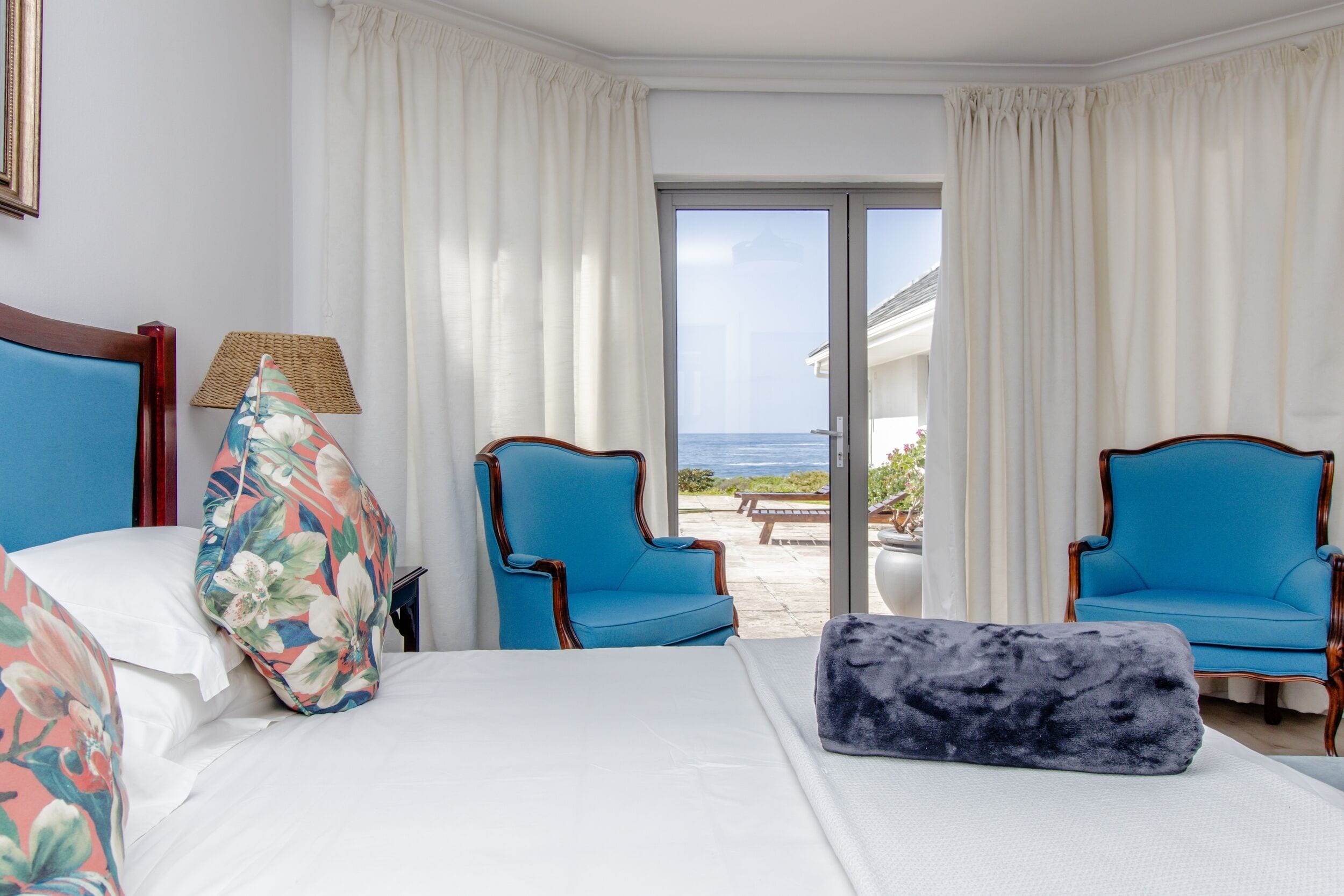 Une chambre d’hôtel avec un lit, deux fauteuils bleus, une vue sur la mer par une porte coulissante et une lampe en osier.
