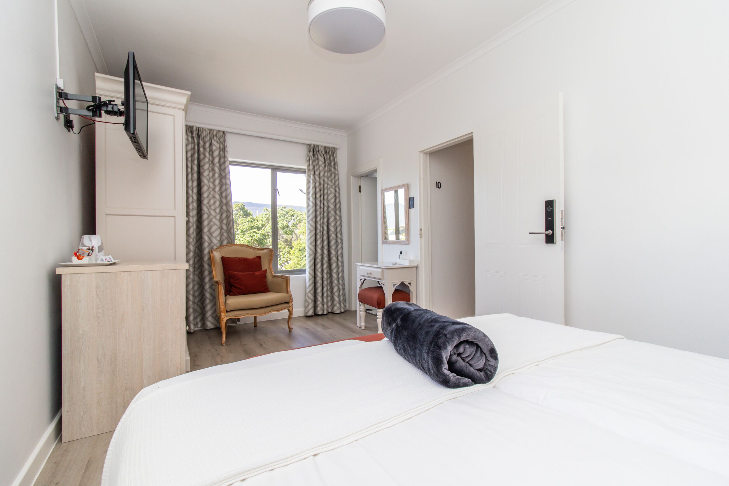 A hotel room with a bed, a desk, a chair, a TV mounted on the wall, and a window with curtains.