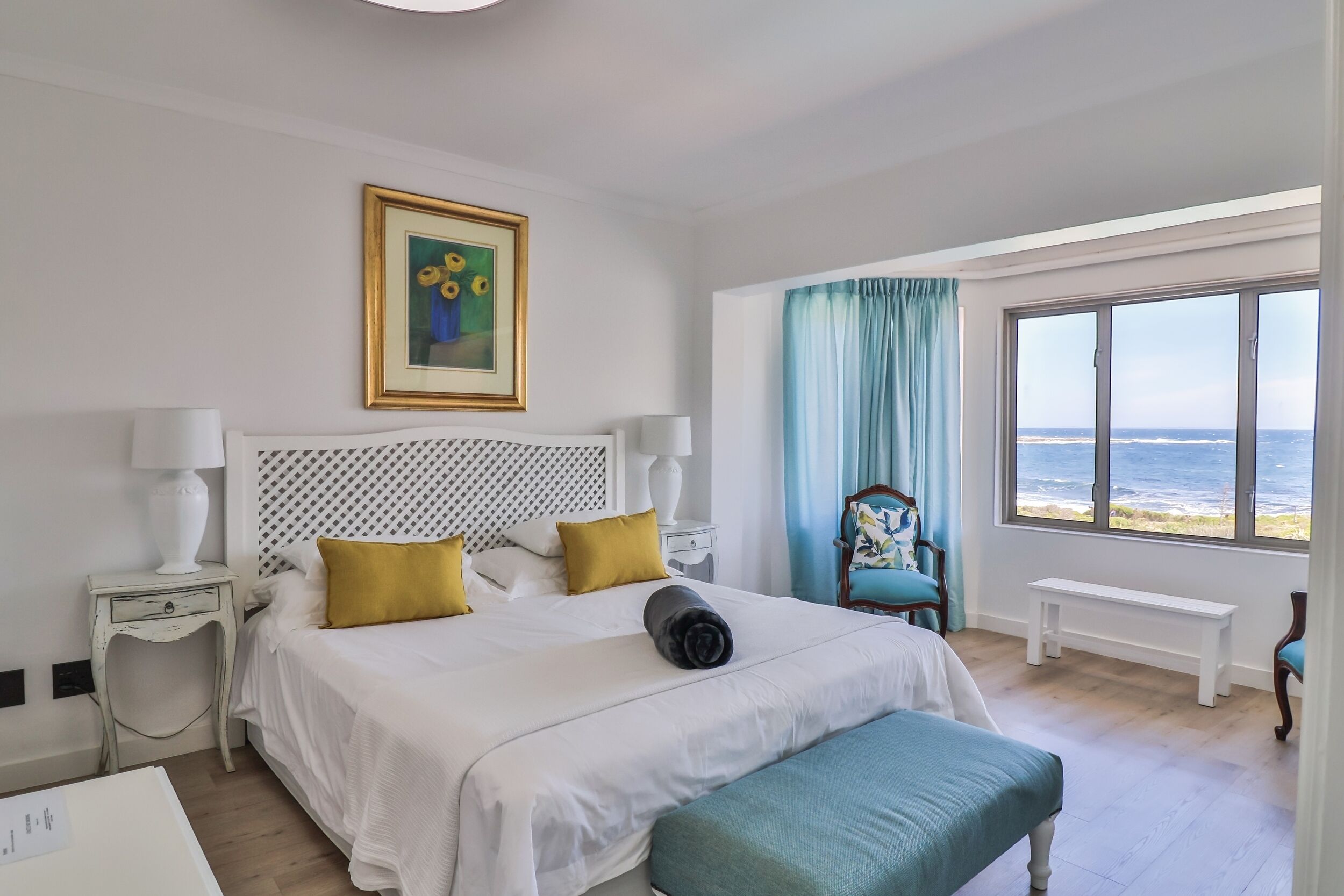 Un cuarto de hotel con una cama, un banco, una silla, una ventana con vista al mar y un cuadro en la pared.