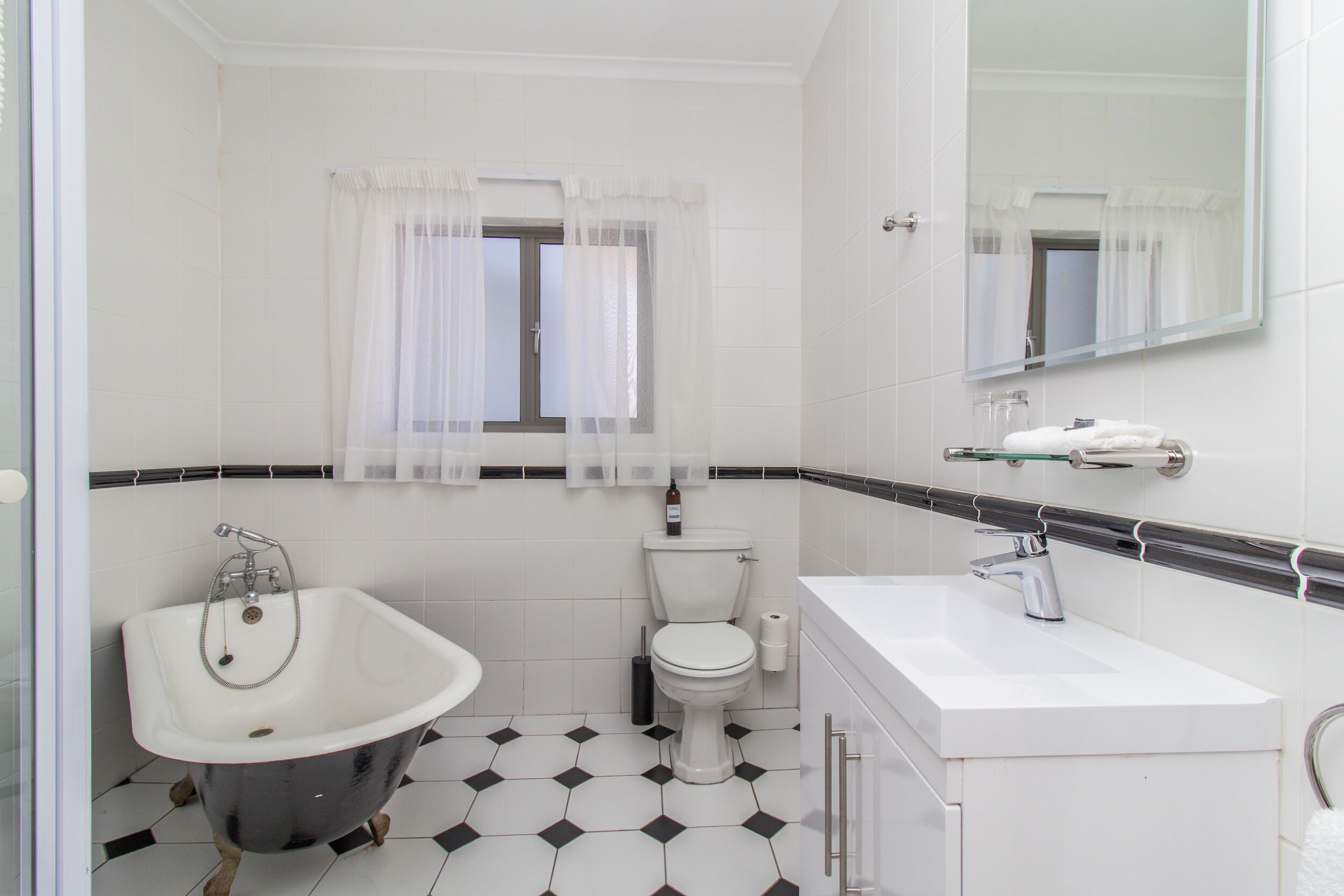 A bathroom with a bathtub, toilet, and sink, featuring a black and white tiled floor and a window with sheer curtains.