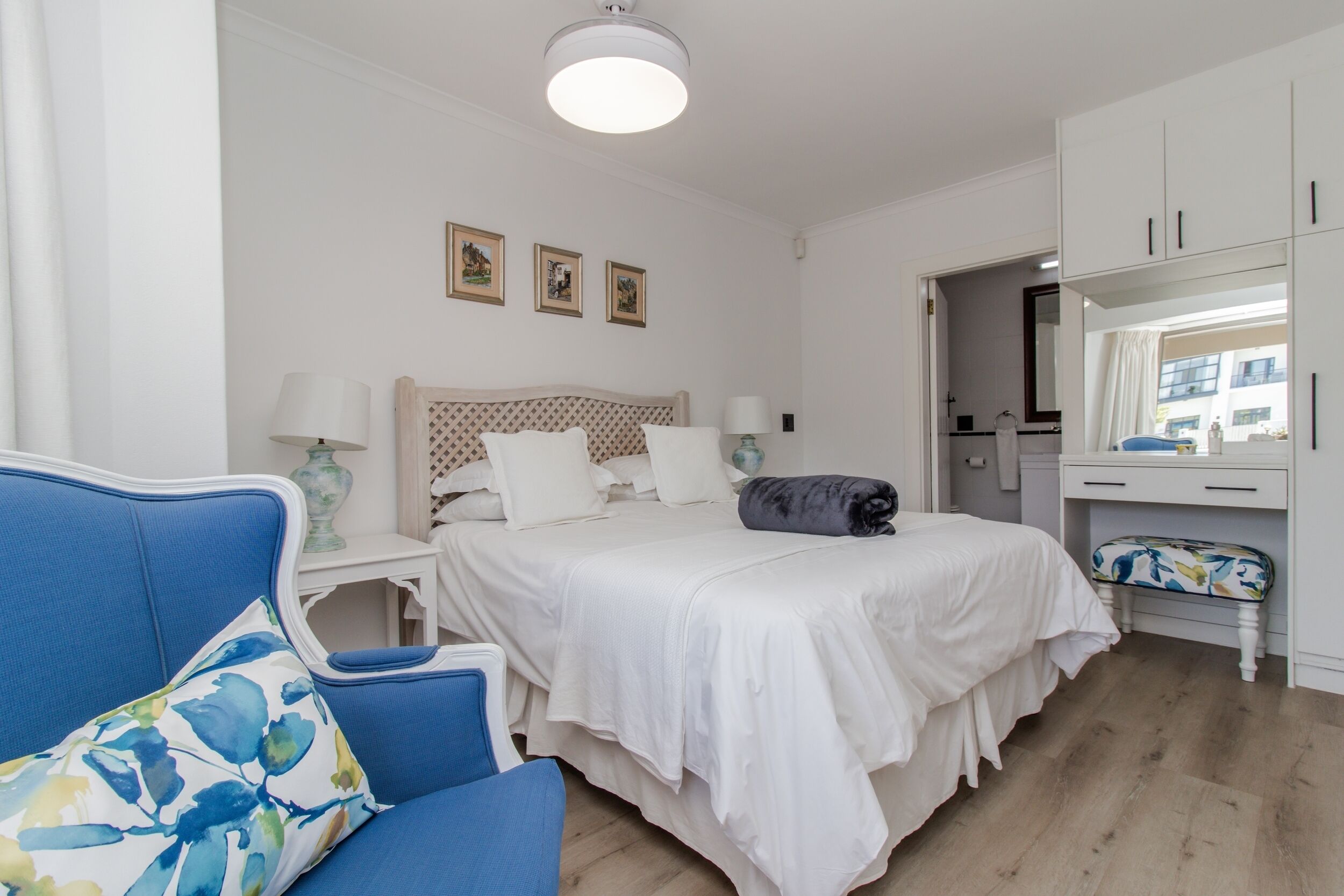 Un dormitorio con una cama, una silla azul, una mesita de noche, un aparador y un espejo.